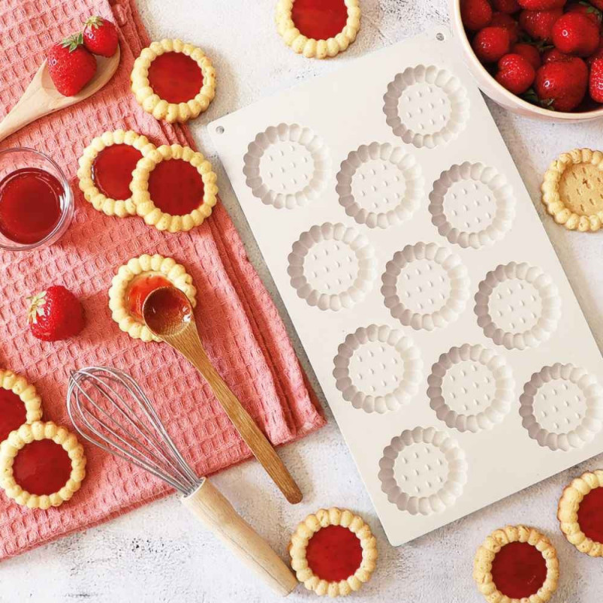 Moule silicone blanc à 12 empreintes pour mini-tartelettes entouré de tartelettes à la fraise, de fraises fraîches et d'ustensiles de pâtisserie.