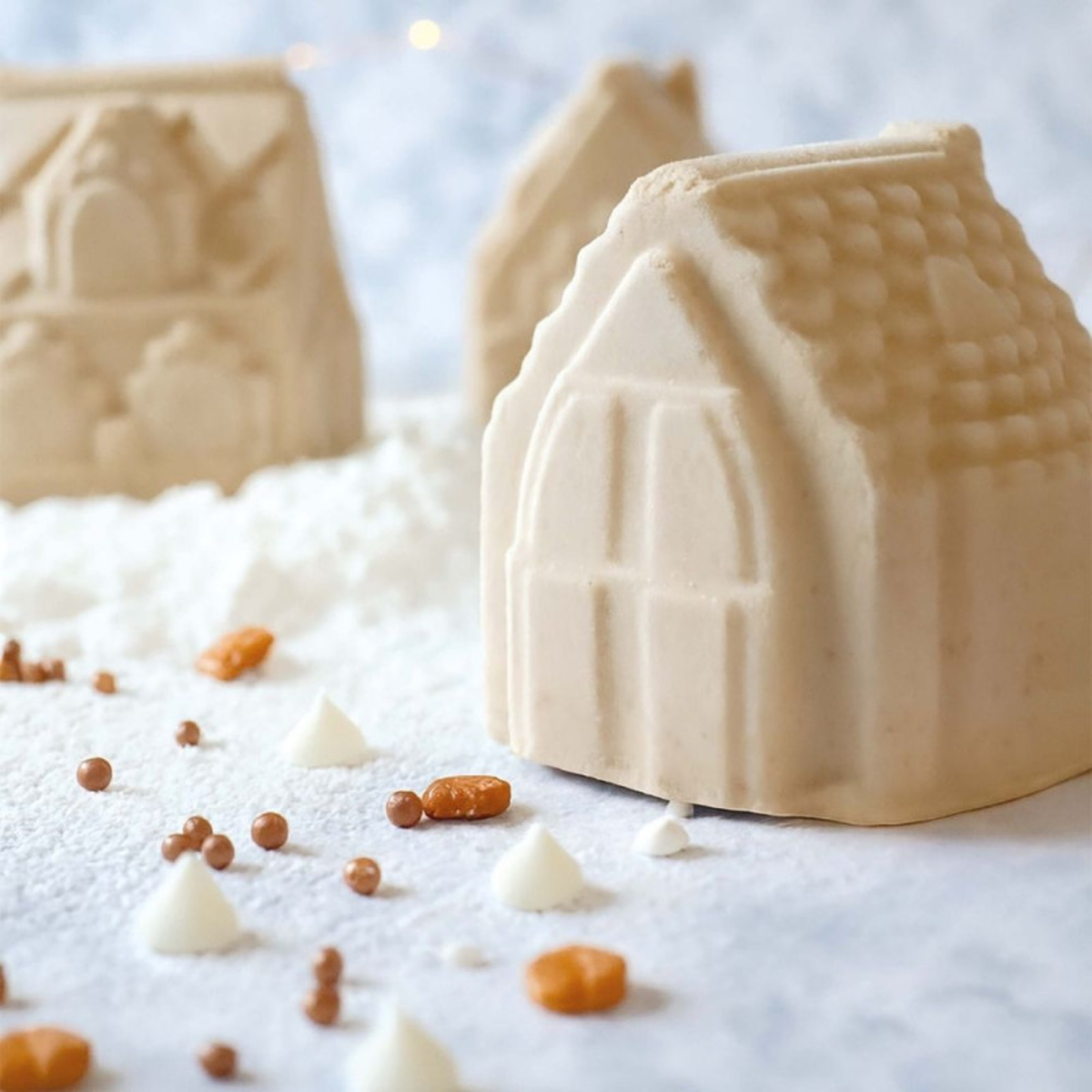 Entremets individuel en forme de chalet de Noël blanc, avec toit en tuiles, porte et fenêtres en relief, présenté dans un décor enneigé.