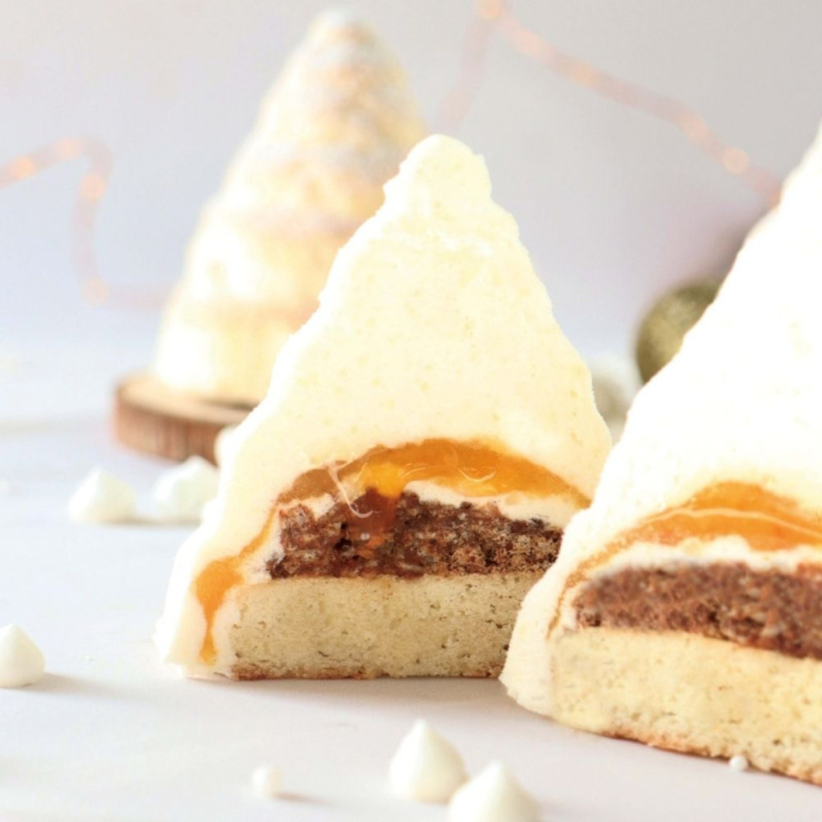 Entremets individuel en forme de sapin de Noël blanc, coupé en deux pour dévoiler un insert orange, un biscuit au chocolat et une mousse claire.