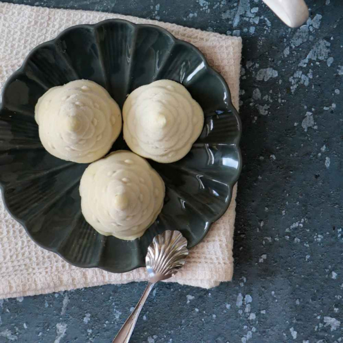 Trois entremets individuels blancs en forme de sapins de Noël 3D, présentés sur une assiette verte foncée.