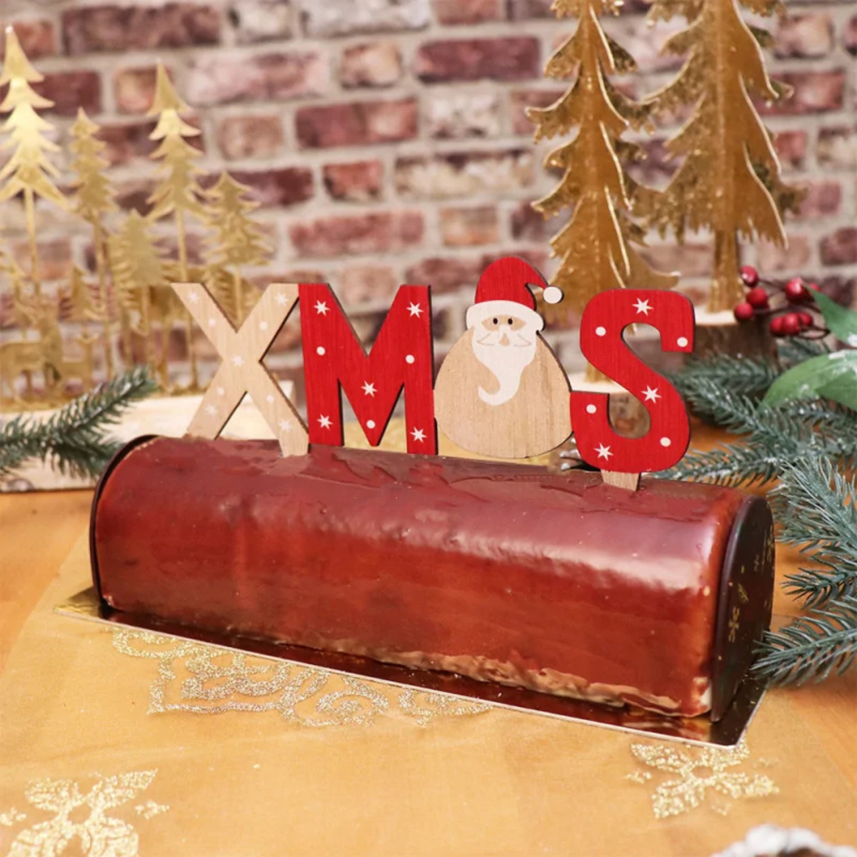 Bûche de Noël au chocolat décorée d'un cake topper Xmas en bois rouge et naturel avec Père Noël, entourée de sapins et de décorations festives.