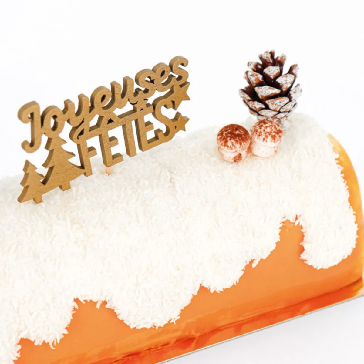Décor à piquer Joyeuses Fêtes doré sur une bûche de Noël blanche à la noix de coco, accompagnée de décorations festives.