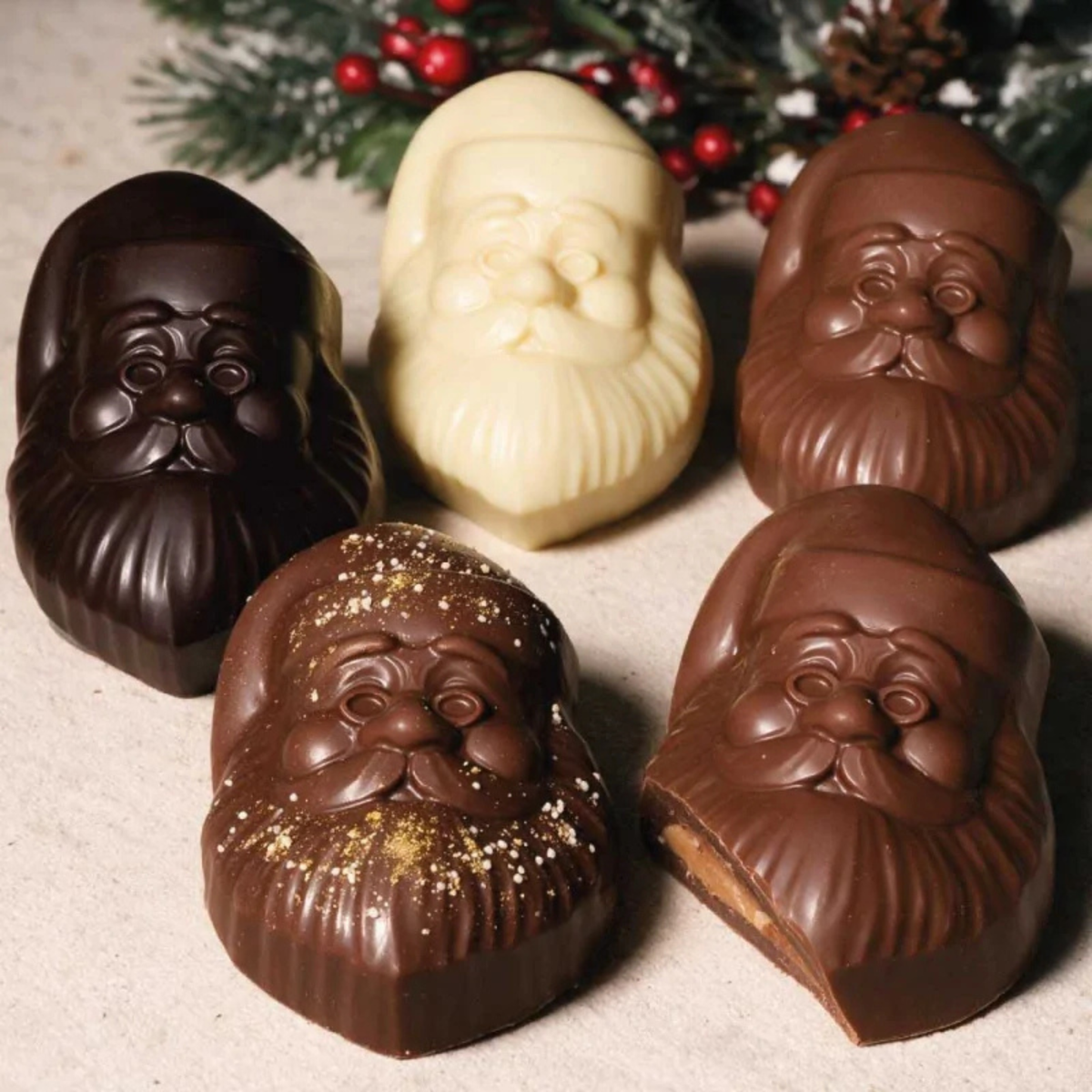 Cinq moulages en chocolat en forme de visage du Père Noël, réalisés en chocolat noir, au lait et blanc, avec détails du bonnet, de la moustache et de la barbe.