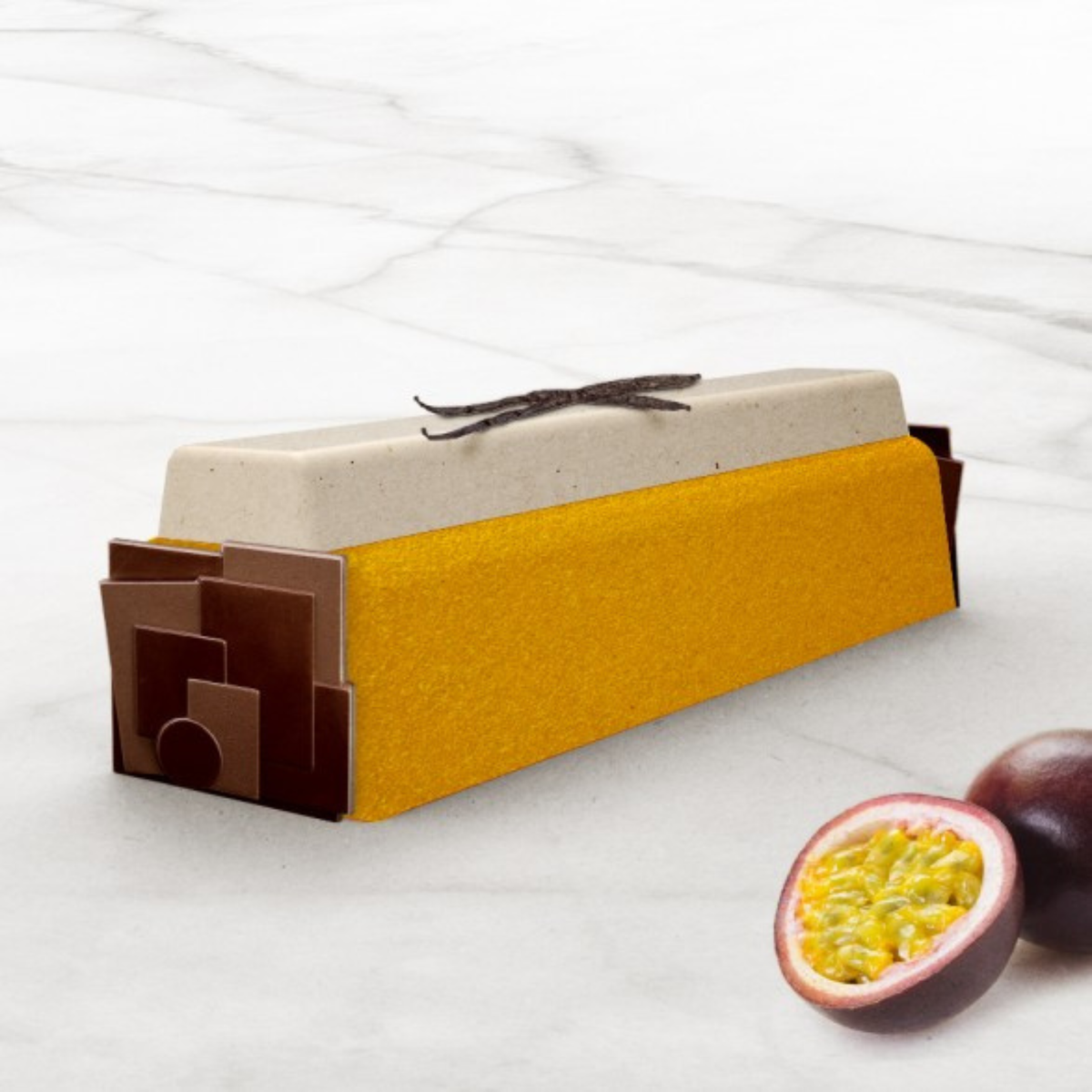 Bûche de Noël rectangulaire avec glaçage doré et mousse vanille, décorée de plaques de chocolat noir et de gousses de vanille, accompagnée de fruits de la passion.