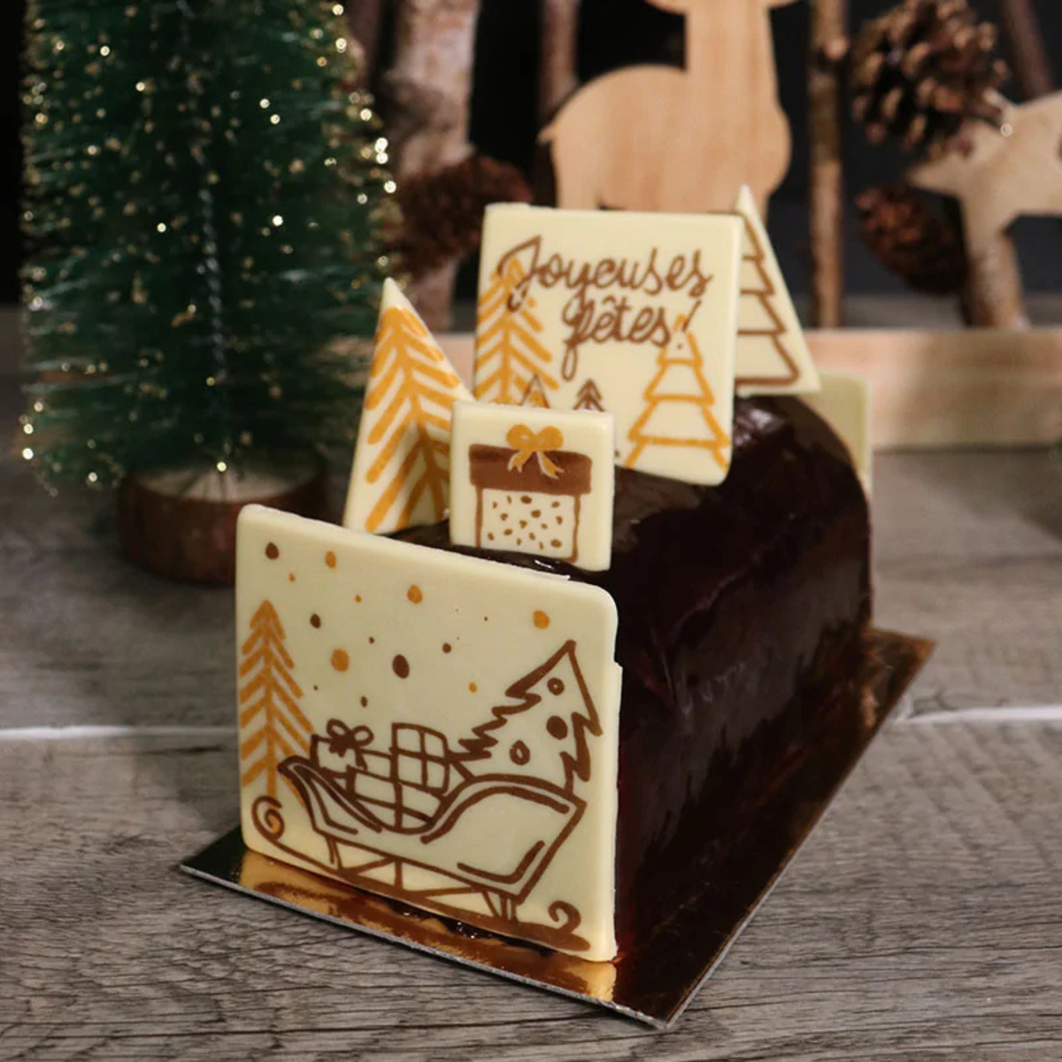 Bûche de Noël au chocolat noir brillant, décorée de plaques en chocolat blanc avec motifs dorés de sapins, traîneau, cadeaux et inscription Joyeuses Fêtes.